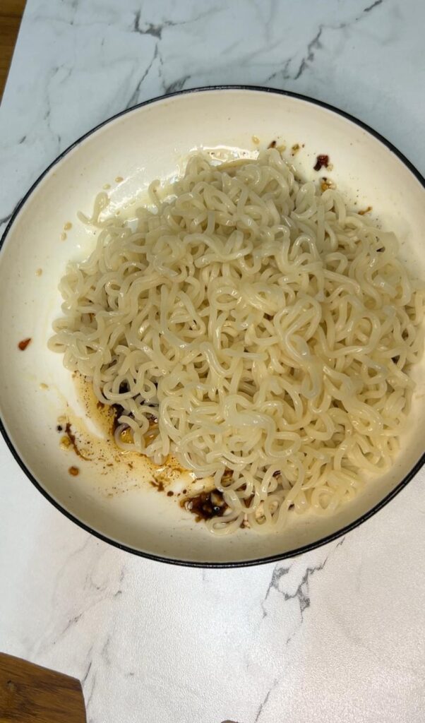 Close-up of chili oil noodles garnished with green onions, sesame seeds, and red chili flakes in a white bowl.